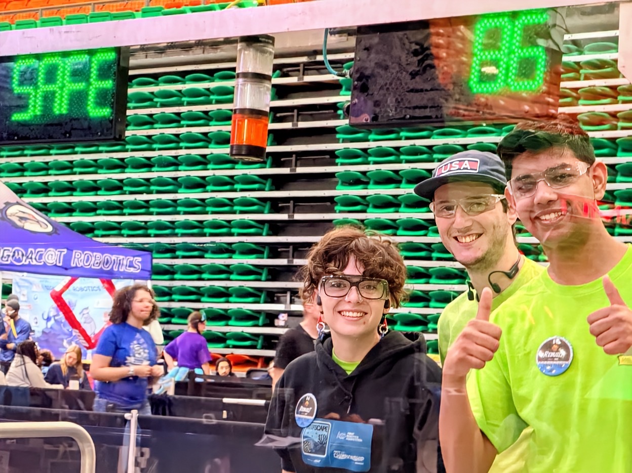 Three students giving a thumbs up in front of the stands, arena scoring displays behind them